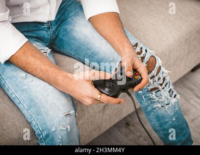 Gay jouant à un jeu d'ordinateur.Un jeune homme en Jean tient le joystick de la console de jeu assis sur le canapé.Gros plan des mains masculines.Entertai Banque D'Images