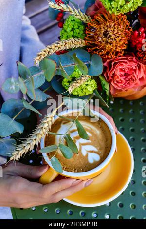 Cappuccino dans une tasse jaune dans les mains de femme avec la composition de fleurs d'automne en citrouille sur une table. Banque D'Images