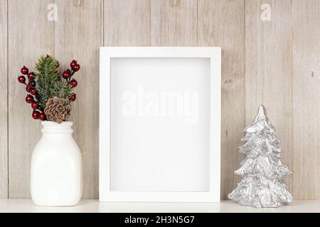 Maquette de cadre blanc avec branches de Noël et arbre argenté sur une étagère.Cadre portrait contre un mur rustique en bois gris. Banque D'Images