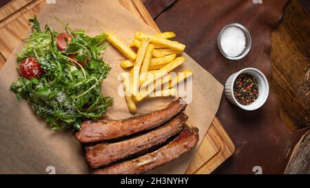 Côte de bœuf avec frites et salade d'arugula avec tomates cerises et parmesan servis à bord recouverts de pa de qualité alimentaire Banque D'Images