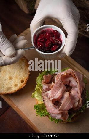 Le chef cuisinier doit faire cuire dans des gants jetables, ajouter la sauce aux cerises à un hamburger de bœuf frais avec des tranches de bacon frites et de laitue fraîche avant Banque D'Images