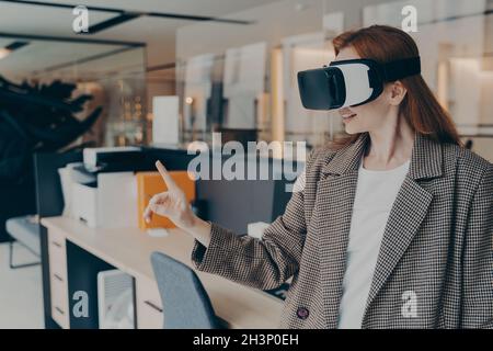 Femme d'affaires souriante et heureuse portant un casque VR, essayant la réalité virtuelle pour la première fois au travail Banque D'Images