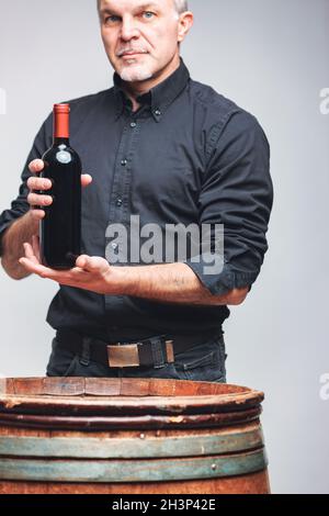 Homme âgé présentant une bouteille de vin rouge non étiquetée dans ses mains alors qu'il se tient derrière un vieux tonneau de maturation en chêne ou un fût dans un portrait de récolte Banque D'Images