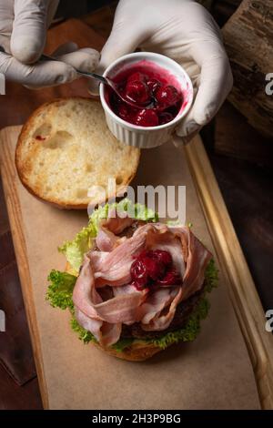 Vue de dessus.Le chef sert un hamburger de bœuf artisanal frais avec des tranches de bacon frites, recouvert de sauce aigre-douce aux cerises et de jambon frais Banque D'Images