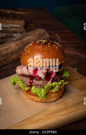 Hamburger artisanal avec sauce aux cerises, tranches de bacon frites, laitue fraîche à côté des lodges en bois décoratifs.Plat servi sur un bois Banque D'Images