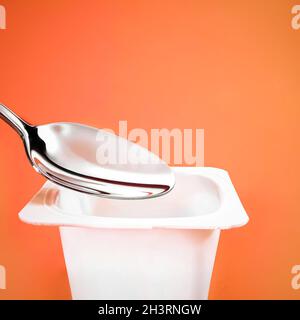 Gobelet à yaourt et cuillère argentée sur fond orange, contenant en plastique blanc avec crème au yaourt, produit laitier frais pour les di sains Banque D'Images