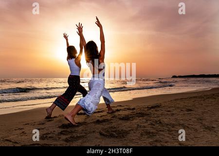 Deux jeunes femmes faisant le guerrier une pose de yoga sur la plage au coucher du soleil.Un sentiment d'énergie et d'amitié dans une atmosphère de fin d'été Banque D'Images