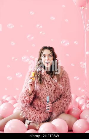 Jeune jolie femme s'amusant avec des bulles de savon sur fond rose avec des ballons Banque D'Images