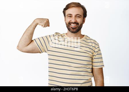 Souriant homme heureux et en bonne santé montrant ses muscles, fléchissant biceps sur le bras, se sentant fort, debout sur fond blanc Banque D'Images