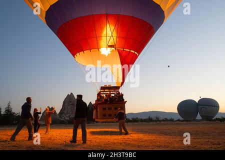GÖREME, TURQUIE - 3 AOÛT 2021: Position des gens pour enlever un panier de ballon d'air chaud plein de touristes tandis que le capitaine allume la flamme du brûleur an Banque D'Images