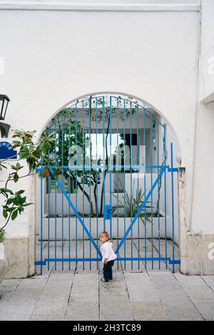 L'enfant se tient dans la cour de la maison et tient sa main à la grille de treillis Banque D'Images