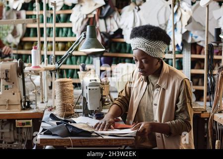 Couturière africaine travaillant avec des motifs et des morceaux de tissus pour coudre de nouveaux vêtements à la table dans l'usine Banque D'Images