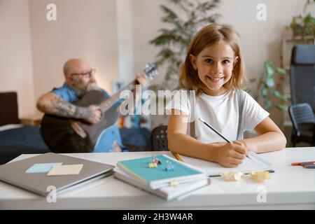 Une petite fille souriante fait ses devoirs tandis que son père joue de la guitare dans son salon Banque D'Images