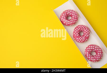 Trois beignets ronds cuits dans un glaçage rose dans une boîte en carton blanc sur fond jaune, vue du dessus Banque D'Images