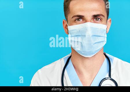 Gros plan portrait d'un beau médecin de sexe masculin portant un masque médical sur fond bleu Banque D'Images