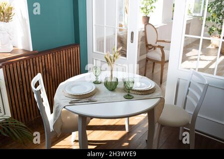 Verres verts et plats blancs sur la table de cuisine, avec plante verte sur le salon en arrière-plan. Banque D'Images