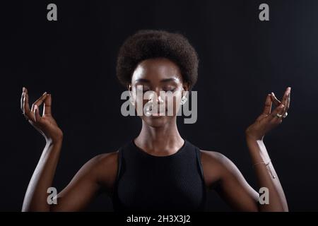 Portrait de studio de la femme afro-américaine montrant un geste zen ou OK.Femme méditant sur fond noir. Banque D'Images