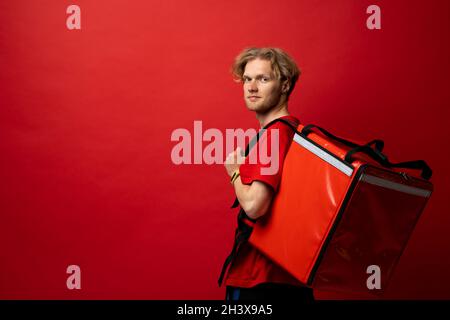 Jeune coursier avec sac thermique sur fond rouge, espace pour le texte.Service de livraison de nourriture.Le livreur dans un t-shirt rouge uniforme travail comme coursier et Banque D'Images