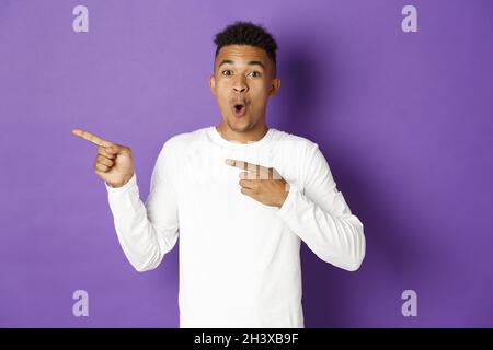 Portrait d'un homme afro-américain émerveillé en sweat-shirt blanc, en regardant impressionné et montrant la publicité, pointant les doigts vers la gauche, Banque D'Images