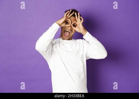 Image d'un homme afro-américain drôle et heureux en sweat-shirt blanc, grimaquant et faisant des visages, debout sur fond violet Banque D'Images