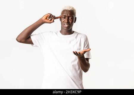 Portrait d'un homme blond afro-américain agacé, se plaignant et collant quelqu'un qui agit stupide, pointant vers la tête, se tenant blanc Banque D'Images