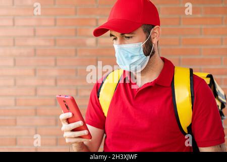 Livraison jeune homme en homme de protection portant un uniforme rouge à l'aide d'un smartphone Banque D'Images