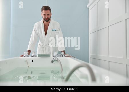 Un homme gai en peignoir doux se penche sur le bain hydromassant et l'eau chaude dans le salon de spa Banque D'Images