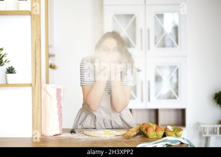 Une jeune femme au foyer en tablier soufflant de la farine tout en faisant de la pâte et de la cuisine dans la cuisine légère à la maison Banque D'Images