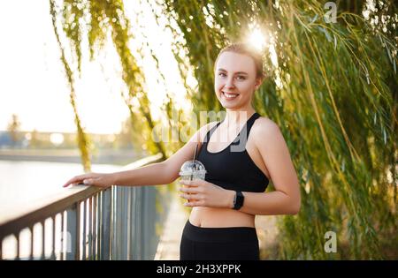 Jeune femme sportive en tenue de sport tenant la limonade à la main et souriant à la caméra tout en se tenant dans le parc près du lac Banque D'Images