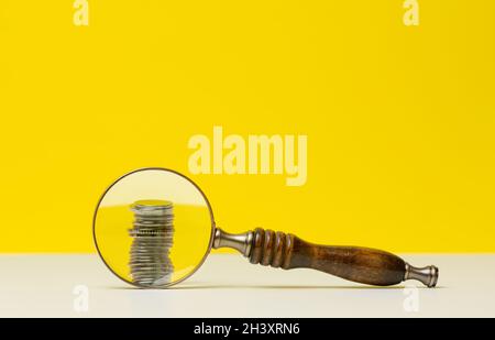 Pile de pièces de monnaie et loupe en bois sur une table blanche, fond jaune.Le concept d'analyse des fonds budgétaires, le contrôle de e Banque D'Images