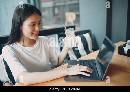 Une jeune femme asiatique boit du soda à la limonade et utilise un ordinateur portable au café en plein air Banque D'Images