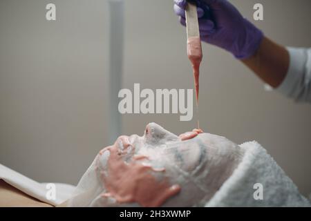 Alginate masque processus d'application. Une femelle mains d'une spatule de maintien d'esthéticienne. Femme couché sur un canapé dans le bureau de cosme Banque D'Images