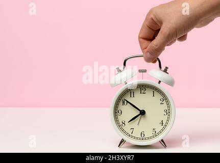 La main féminine tient un réveil en métal blanc sur une table blanche, le temps est de dix à huit le matin Banque D'Images