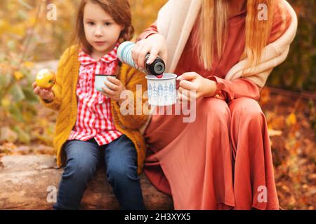 Jeune famille, mère et petite fille, dans des vêtements élégants appréciant une boisson chaude dans la forêt d'automne Banque D'Images
