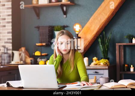 Jeune femme pensive pigiste assis à la table à la maison avec ordinateur portable, prenant des notes Banque D'Images