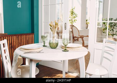 Verres verts et plats blancs sur la table ronde de la cuisine, avec plante verte sur le salon en arrière-plan. Banque D'Images