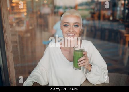 Jeune femme blonde millénial cheveux courts intérieur avec smoothie vert Banque D'Images