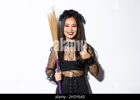 Image d'une femme asiatique souriante en costume de sorcière avec un balai, montrant le pouce en approbation, debout sur fond blanc Banque D'Images