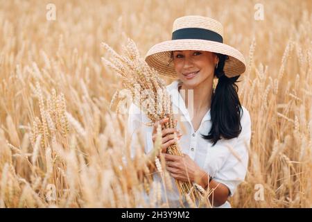 Jeune femme en chapeau de paille tenant une feuille d'épis de blé au champ agricole Banque D'Images