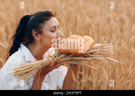 Femme tenant et sentant du pain de blé fait maison et de la Gerbe d'oreilles dans les mains dans le champ de blé au coucher du soleil. Banque D'Images