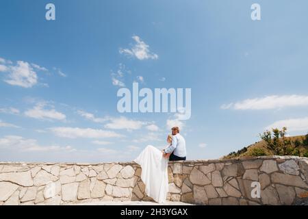 La mariée et le marié sont assis sur une clôture en pierre, la mariée est penchée sur le marié, le marié la prend doucement Banque D'Images