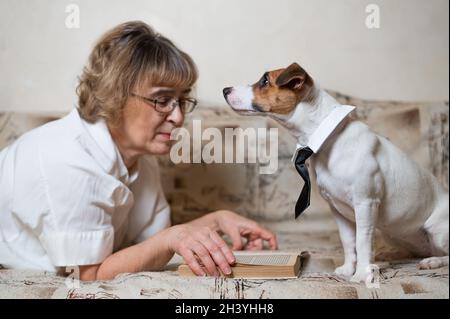 Une femme caucasienne âgée est allongée sur un canapé avec un chien intelligent Jack russell terrier portant des lunettes et une cravate et lisant un livre. Banque D'Images