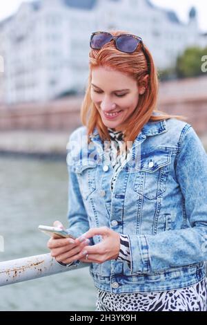 Bonne jeune femme caucasienne à tête rouge lisant un message sur un smartphone à la banque de la rivière Banque D'Images