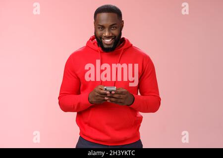 Attrayant heureux souriant afro-américain barbu gars dans rouge hoodie jouant à des jeux smartphone grinning joyeusement messagerie ayant Banque D'Images