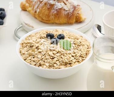 Flocons d'avoine crus dans une assiette en céramique blanche sur une table blanche, petit déjeuner Banque D'Images