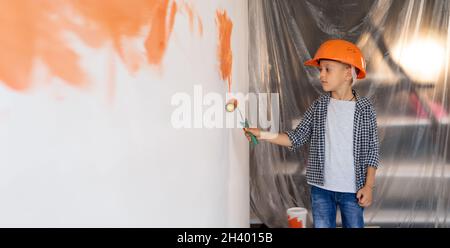 les enfants peignent à l'intérieur.Un petit garçon dans un casque orange répare une maison peint le mur avec un rouleau.L'enfant aide à décorer les murs. Rénovation Banque D'Images