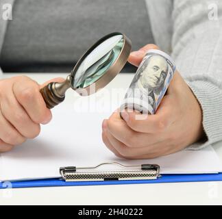 Une pile de cent billets de dollars dans la main d'une femme et une loupe en bois sur une table blanche.Analyse budgétaire, revenu et Banque D'Images