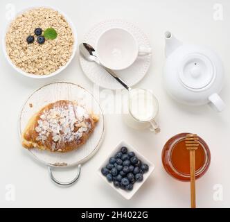 Petit déjeuner du matin, flocons d'avoine crus dans une assiette en céramique, lait dans un carafe, bleuets et miel dans un pot sur une table blanche Banque D'Images