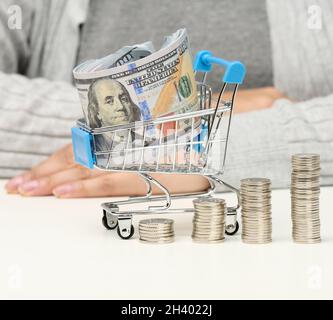 Femme assis à une table, des piles de pièces de monnaie blanches et un chariot miniature avec des dollars américains.Concept d'épargne, de vente, de taxe Banque D'Images