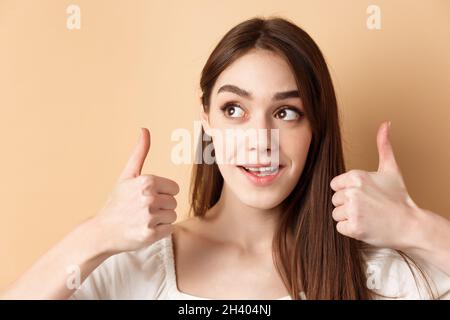 Gros plan de la jolie fille regarder le logo du coin supérieur gauche, montrant les pouces vers le haut dans l'approbation, comme le produit, debout sur le fond beige Banque D'Images